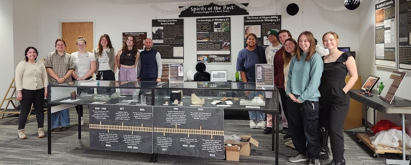 Smiling students standing in front of Spirits of the Past: Archaeology of a Ghost Town, an exhibit about Winnipeg Junction displayed at the AU library