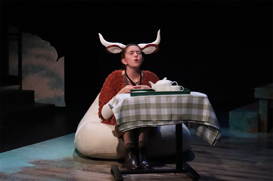 A character from Sometimes the Rain, Sometimes the Sea seated at a table with a teapot, wearing a cow's ears and horns.