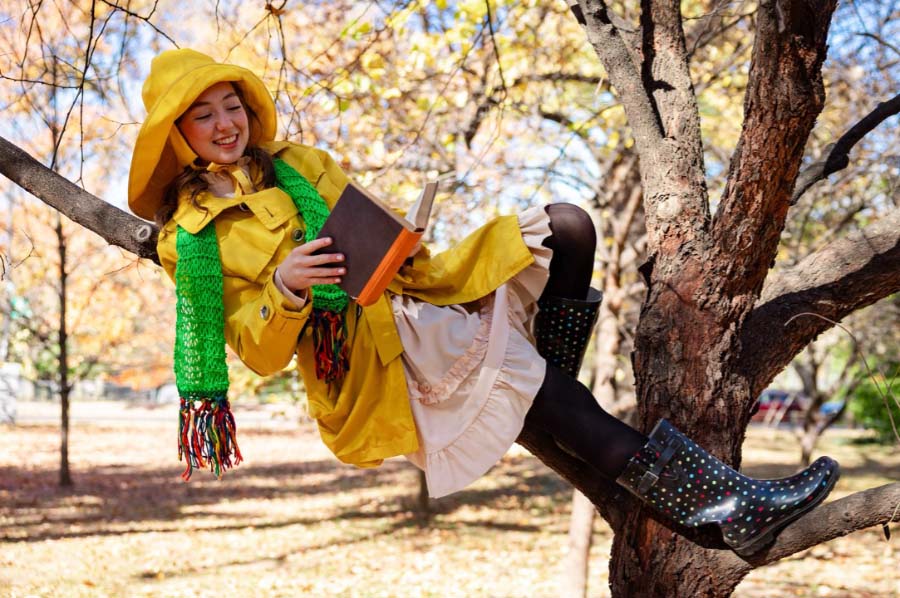 A character from Sometimes the Rain, Sometimes the Sea wearing a yellow raincoat and rain hat, green scarf, pink dress and multicolored rainboots