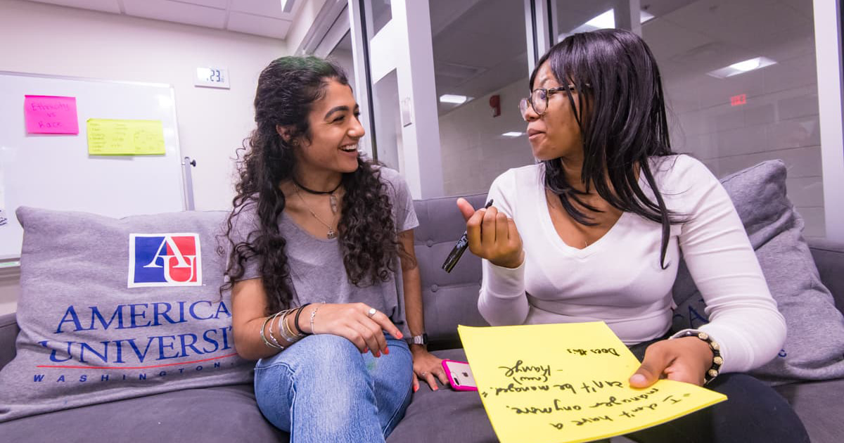 Two students sit on a couch as they have a conversation.