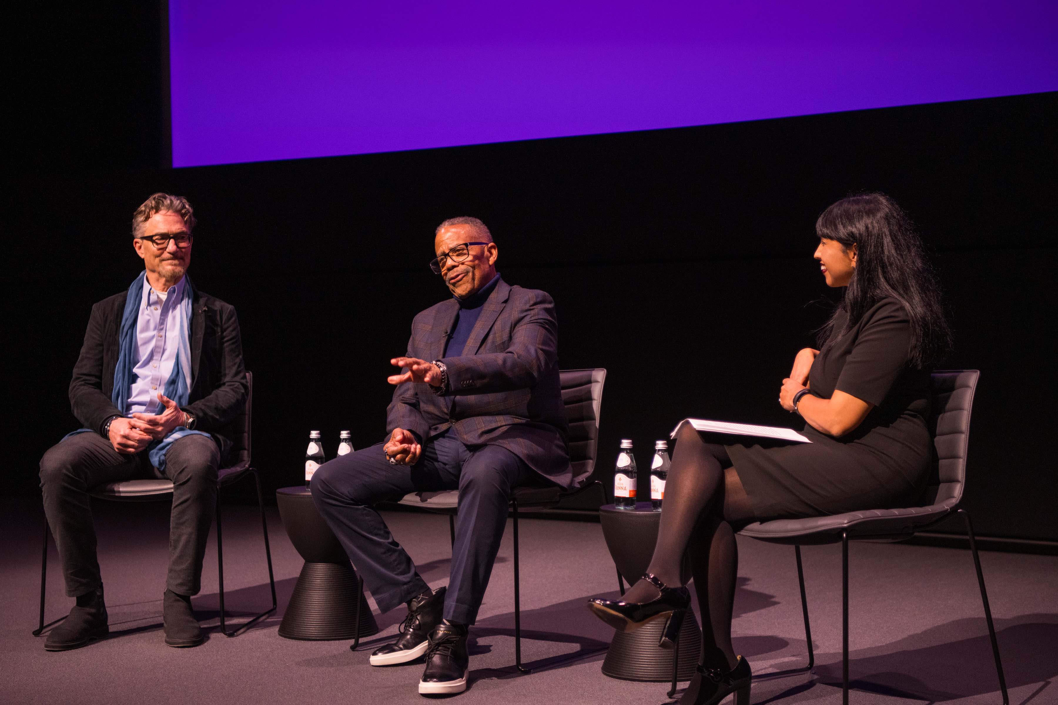 Russell Williams II, SOC/BA ’74, and Barry Josephson, SPA/BA ’78, on stage with  SOC Interim Dean Leena Jayaswal at the Academy Museum of Motion Pictures. Images from Williams’ credits, including Field of Dreams, are displayed on the theater screen.