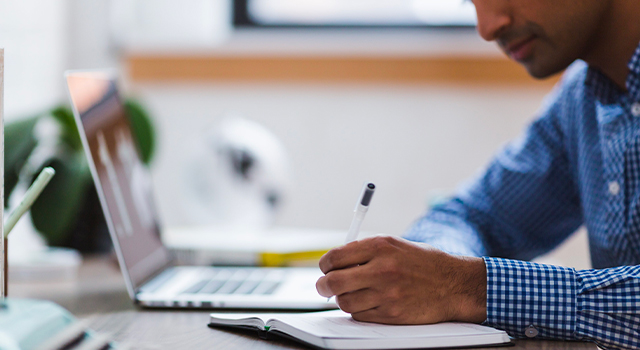 man writes with a pen in a journal next to his laptop at a brightly lit desk