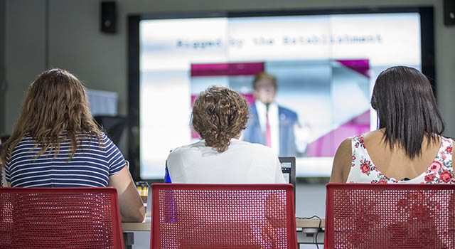 The backs of three students watching a video of president Trump in a class.