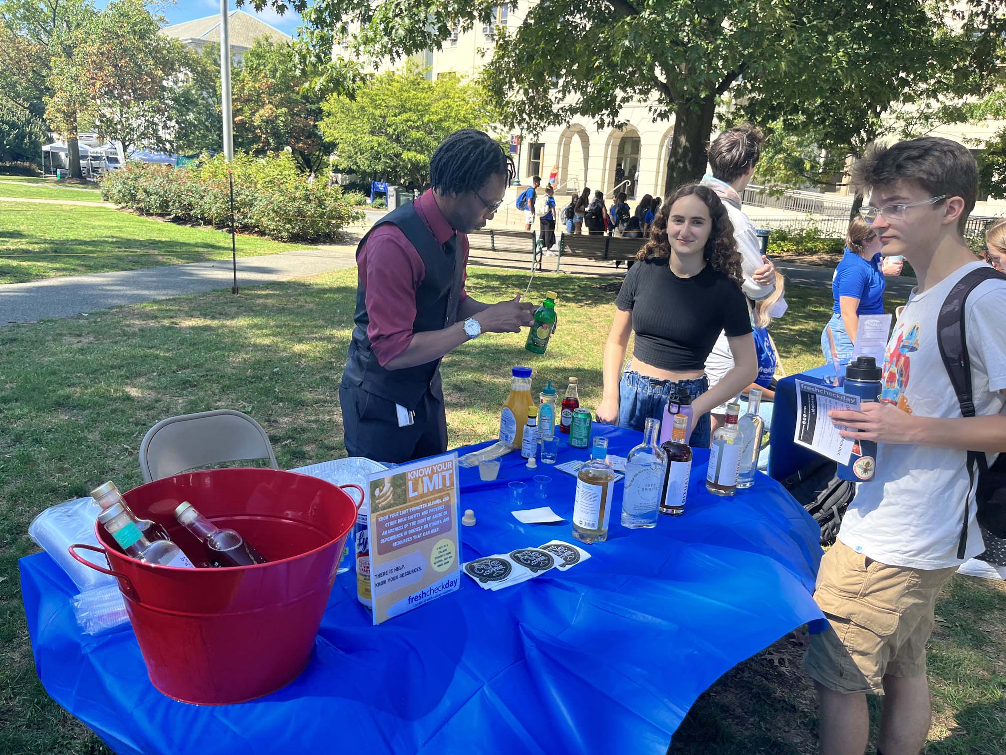 A table for Fresh Check Day has three people gathered around, with one using several bottles on the table to make mocktails.