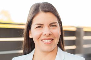 Woman in sport jacket, standing outside in front of a fence.