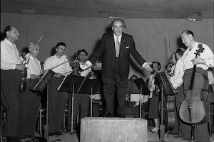 Man in tuxedo stands on podium surrounded by orchestra members holding instruments