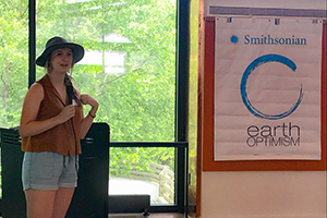 Beth Ebisch standing in front of a window holding a microphone and speaking